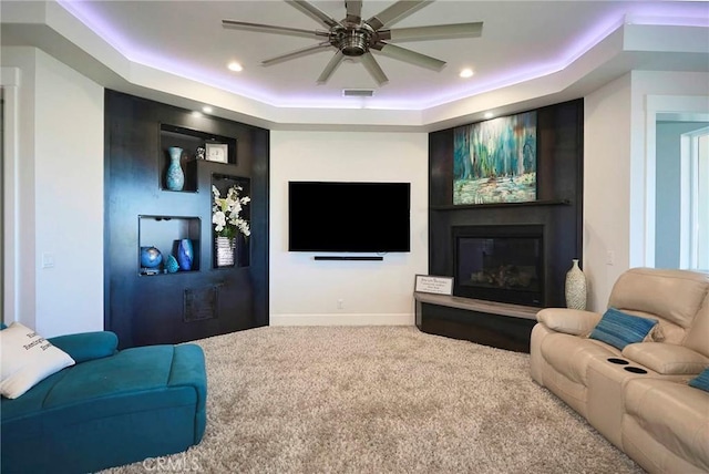 living area featuring carpet floors, visible vents, a glass covered fireplace, a tray ceiling, and a ceiling fan