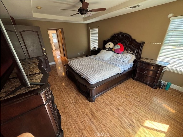 bedroom featuring ceiling fan, visible vents, baseboards, a tray ceiling, and light wood-style flooring