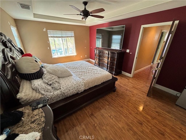 bedroom with ceiling fan, baseboards, wood finished floors, a tray ceiling, and visible vents