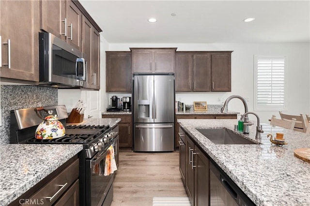 kitchen featuring light stone countertops, dark brown cabinetry, appliances with stainless steel finishes, a sink, and light wood-style floors