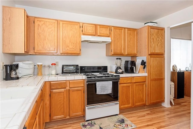 kitchen featuring light wood finished floors, range with gas stovetop, under cabinet range hood, and tile countertops