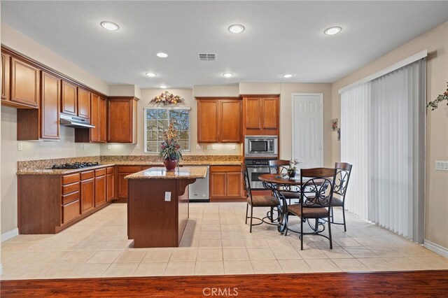 562 Silverhawk Dr, Corona, CA 92879 - kitchen featuring appliances with stainless steel finishes, under cabinet range hood, brown cabinetry, light stone countertops, and a kitchen island