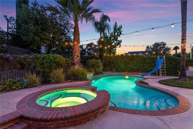 pool at dusk featuring a fenced backyard, a water slide, and a pool with connected hot tub