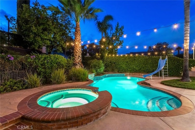 pool at twilight with a patio area, a water slide, and a pool with connected hot tub