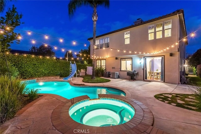 back of house at night with a pool with connected hot tub, cooling unit, a patio area, and stucco siding