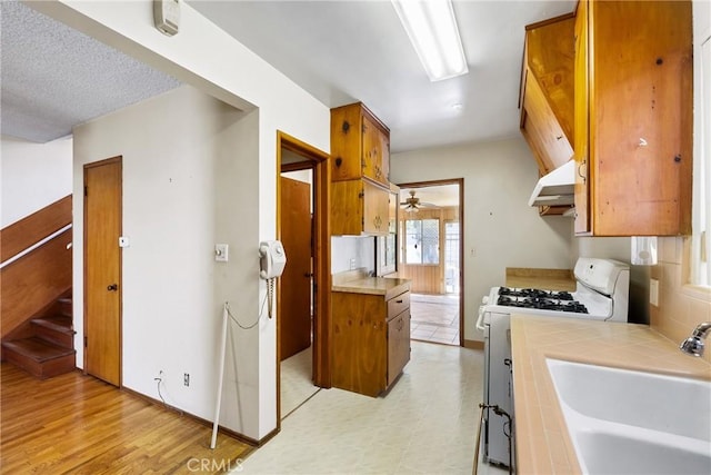 kitchen with brown cabinets, a sink, under cabinet range hood, tile counters, and white range with gas stovetop
