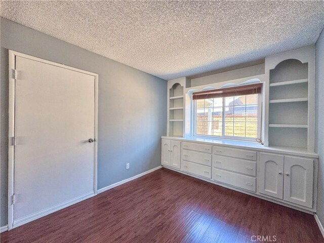 spare room with a textured ceiling, built in shelves, baseboards, and dark wood-type flooring