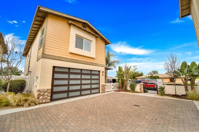 view of side of property featuring a gate, decorative driveway, an attached garage, stone siding, and stucco siding