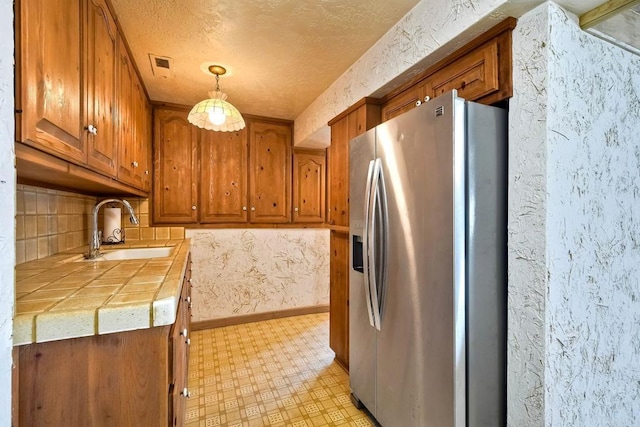 kitchen featuring visible vents, stainless steel fridge, tile countertops, brown cabinets, and a sink