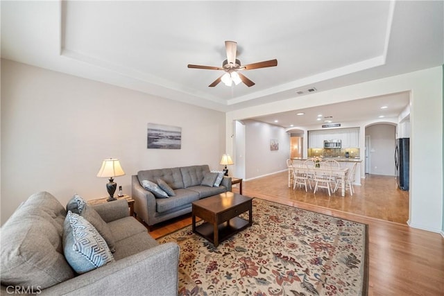living area with a raised ceiling, arched walkways, a ceiling fan, light wood-type flooring, and visible vents