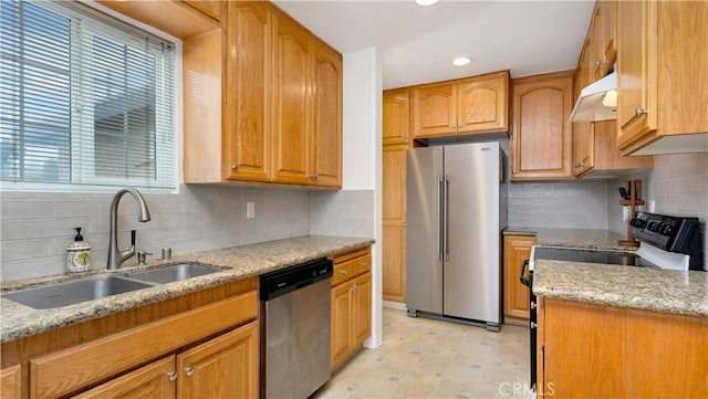 kitchen with appliances with stainless steel finishes, tasteful backsplash, light stone countertops, a sink, and under cabinet range hood