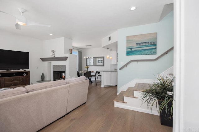 living area with a ceiling fan, wood finished floors, stairway, a warm lit fireplace, and recessed lighting