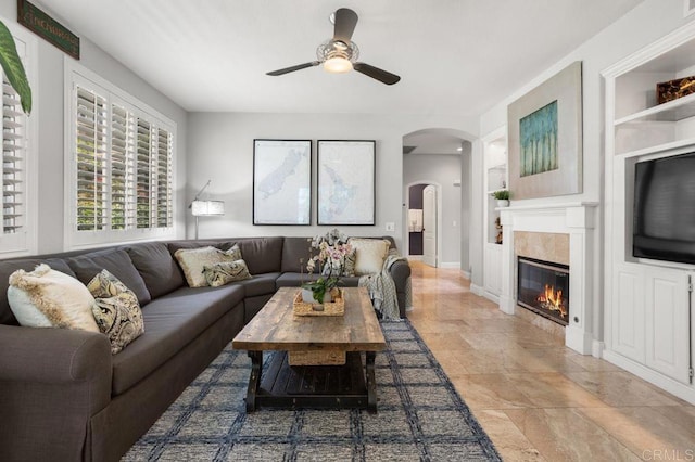 living area with ceiling fan, stone finish flooring, built in features, arched walkways, and a glass covered fireplace
