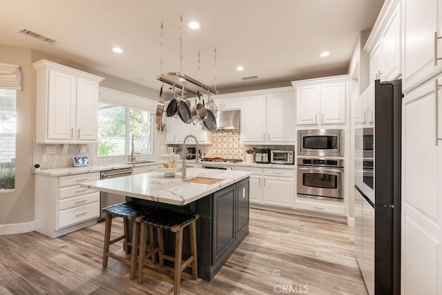 kitchen with wall chimney range hood, appliances with stainless steel finishes, an island with sink, white cabinetry, and visible vents