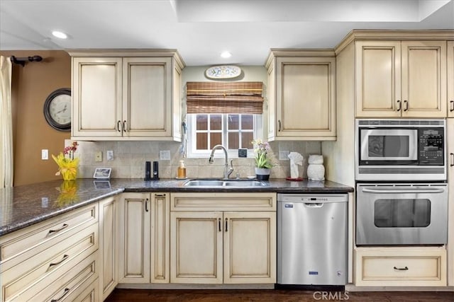 kitchen with dark stone counters, cream cabinets, a sink, appliances with stainless steel finishes, and backsplash