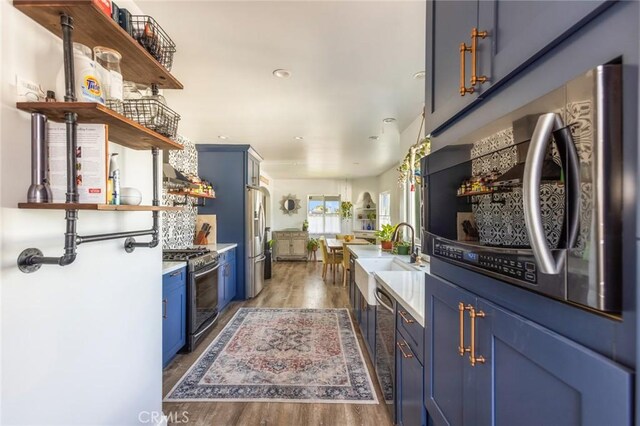 kitchen with blue cabinetry, appliances with stainless steel finishes, open shelves, wood finished floors, and a sink