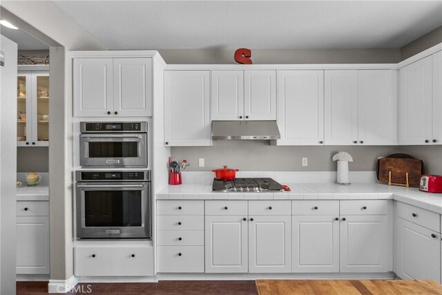 kitchen with tile countertops, white cabinetry, under cabinet range hood, light wood-style flooring, and stainless steel appliances