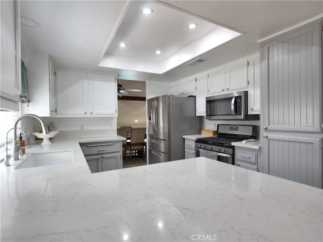 kitchen with light stone counters, a sink, stainless steel appliances, a ceiling fan, and a tray ceiling