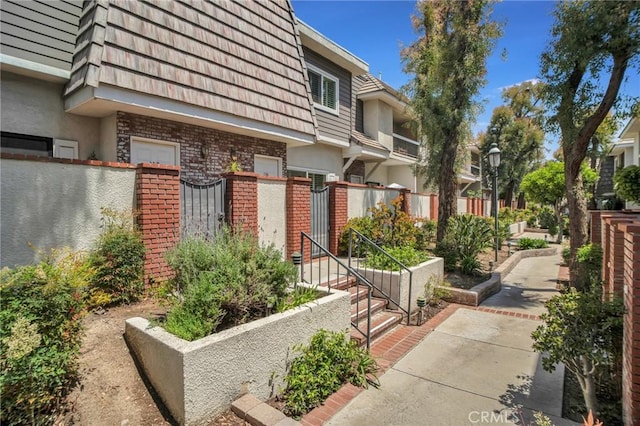 exterior space with a gate, brick siding, mansard roof, fence, and stucco siding
