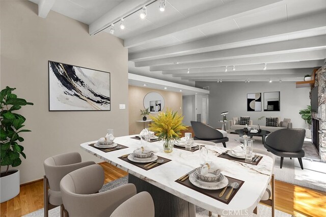 dining area featuring beamed ceiling, track lighting, a stone fireplace, and light wood-type flooring