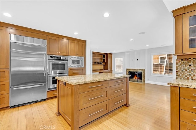 kitchen with a kitchen island, light stone countertops, light wood finished floors, a warm lit fireplace, and built in appliances