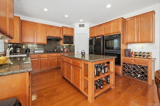 kitchen featuring visible vents, wood finished floors, under cabinet range hood, dark stone countertops, and black appliances