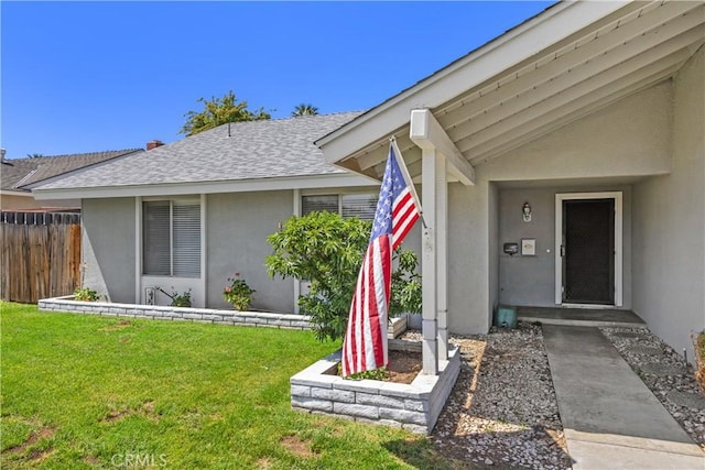 view of exterior entry with a lawn, a shingled roof, stucco siding, and fence