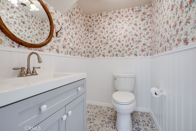 half bathroom featuring vanity, toilet, wallpapered walls, tile patterned floors, and a wainscoted wall