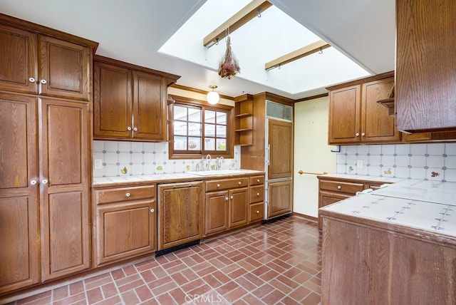 kitchen with brown cabinetry, open shelves, light countertops, brick floor, and paneled dishwasher