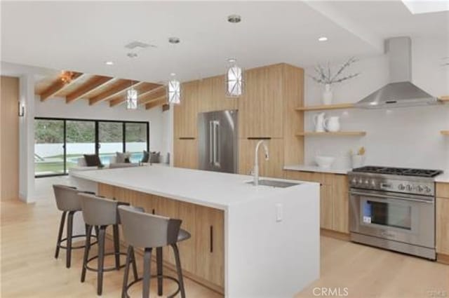 kitchen with wall chimney range hood, modern cabinets, a sink, light brown cabinets, and premium appliances