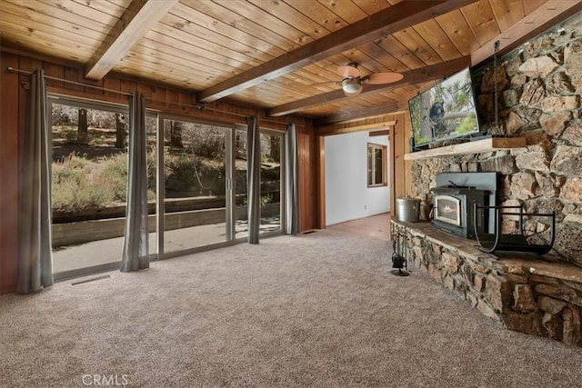 unfurnished living room featuring a wood stove, beamed ceiling, ceiling fan, wood ceiling, and carpet flooring