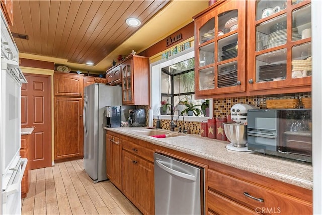 kitchen featuring appliances with stainless steel finishes, a sink, brown cabinetry, backsplash, and glass insert cabinets