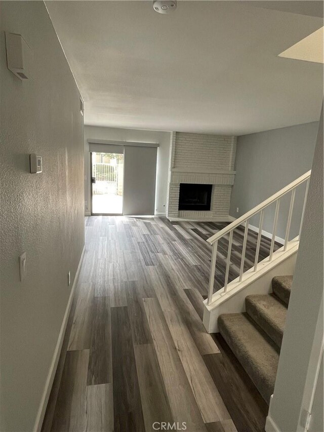 unfurnished living room featuring a brick fireplace, stairs, baseboards, and wood finished floors