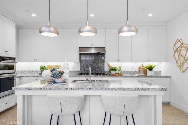 kitchen featuring light stone countertops, a breakfast bar area, an island with sink, white cabinetry, and under cabinet range hood