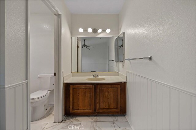 bathroom with a ceiling fan, vanity, a wainscoted wall, toilet, and marble finish floor