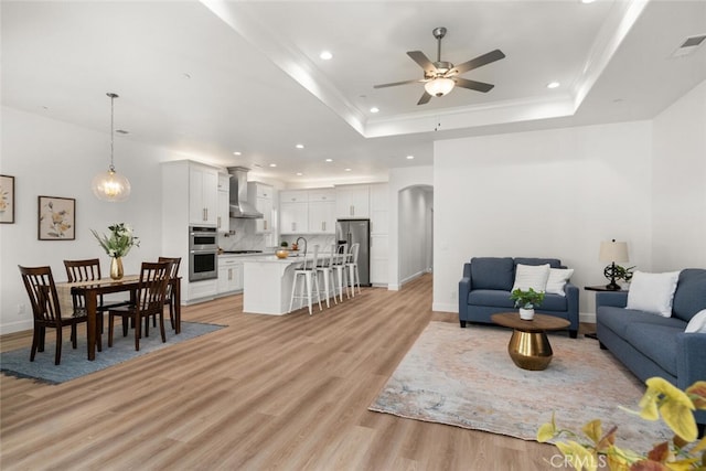living room featuring ceiling fan, light wood finished floors, a raised ceiling, arched walkways, and recessed lighting