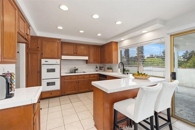 kitchen featuring tile countertops, brown cabinets, white double oven, a sink, and a peninsula