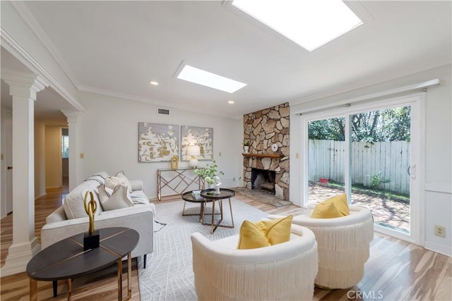 living room with decorative columns, a stone fireplace, crown molding, a skylight, and light wood-type flooring