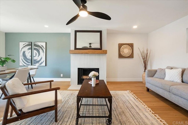 living room with a large fireplace, ceiling fan, baseboards, and light wood-style flooring