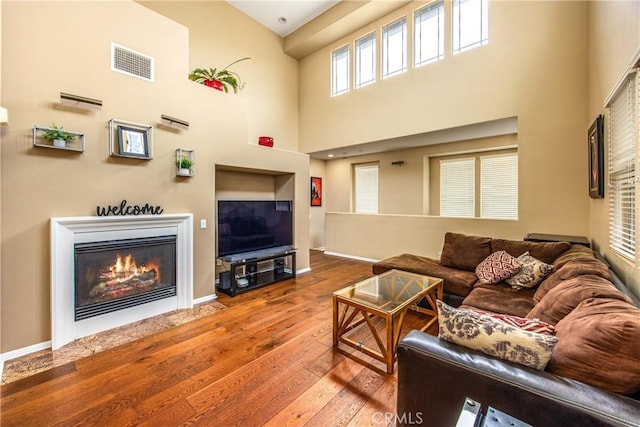living area with visible vents, wood-type flooring, a glass covered fireplace, a towering ceiling, and baseboards