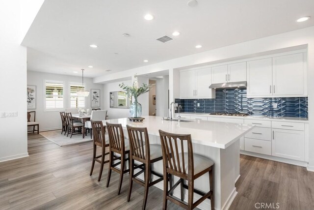 kitchen featuring a sink, light countertops, gas stovetop, under cabinet range hood, and a center island with sink