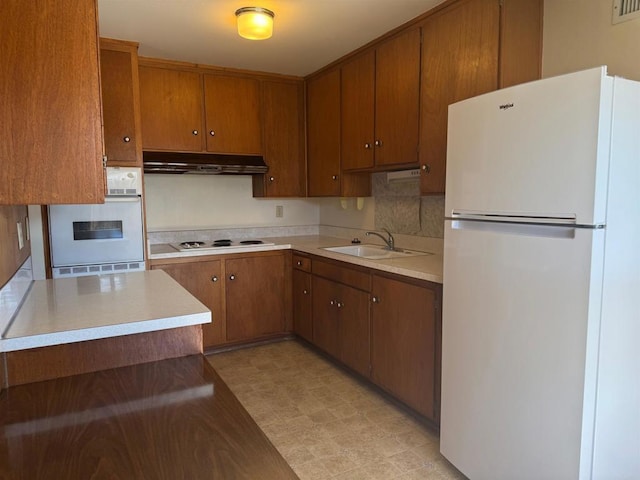 kitchen featuring brown cabinetry, a sink, light floors, under cabinet range hood, and white appliances