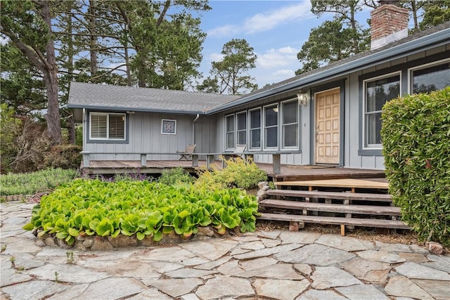 view of exterior entry featuring board and batten siding, a wooden deck, a shingled roof, and a chimney