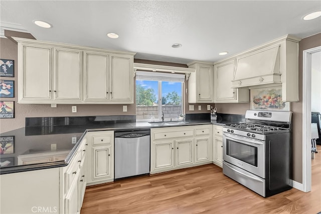 kitchen featuring appliances with stainless steel finishes, dark countertops, a sink, light wood-style floors, and custom range hood