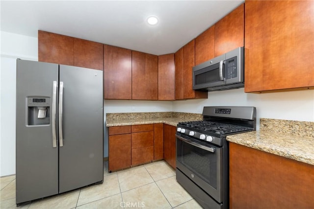 kitchen with appliances with stainless steel finishes, light tile patterned flooring, brown cabinetry, and light stone countertops