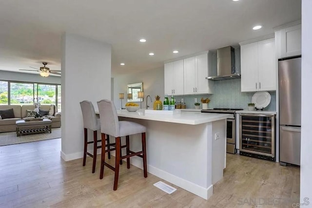 kitchen with beverage cooler, light wood-style floors, wall chimney range hood, and stainless steel appliances