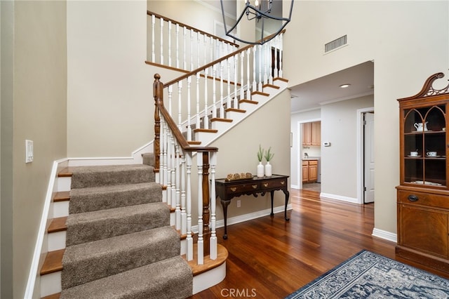 staircase with a towering ceiling, visible vents, wood finished floors, baseboards, and ornamental molding