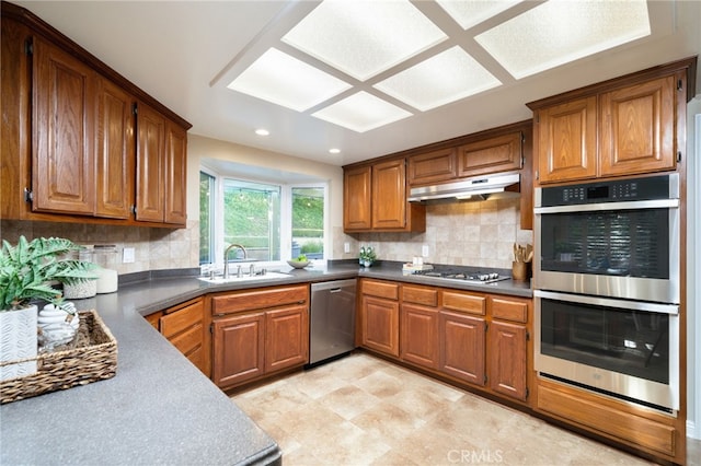 kitchen with brown cabinets, a sink, stainless steel appliances, decorative backsplash, and under cabinet range hood
