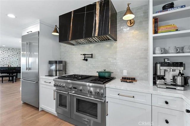 kitchen with light wood-type flooring, wall chimney exhaust hood, premium appliances, decorative backsplash, and white cabinets
