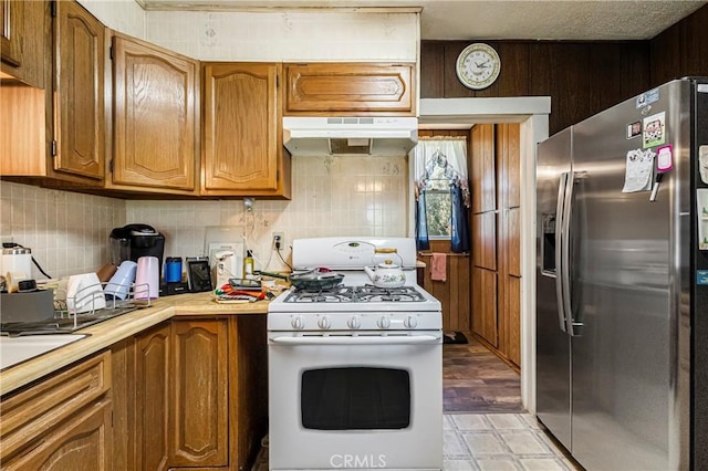 kitchen featuring white gas stove, backsplash, light countertops, stainless steel fridge, and under cabinet range hood